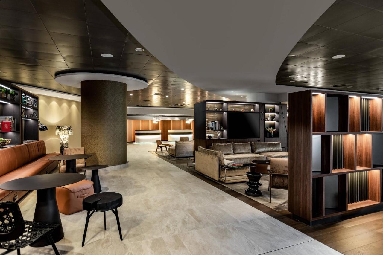 Frankfurt Airport Marriott Hotel فرانكفورت المظهر الخارجي الصورة The photo shows a modern and stylish interior space, likely a hotel lobby or lounge. It features curved walls and a sleek ceiling design. There are comfortable seating areas with a mix of sofas and chairs arranged around small tables. A shelving unit