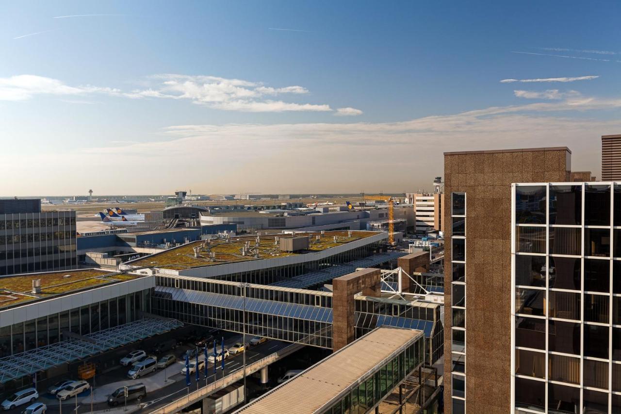 Frankfurt Airport Marriott Hotel فرانكفورت المظهر الخارجي الصورة The photo shows an aerial view of an airport. In the foreground, there are modern buildings and structures, including a glass-fronted building with multiple stories. Surrounding the airport area, there are runways and taxiways visible, along with a v
