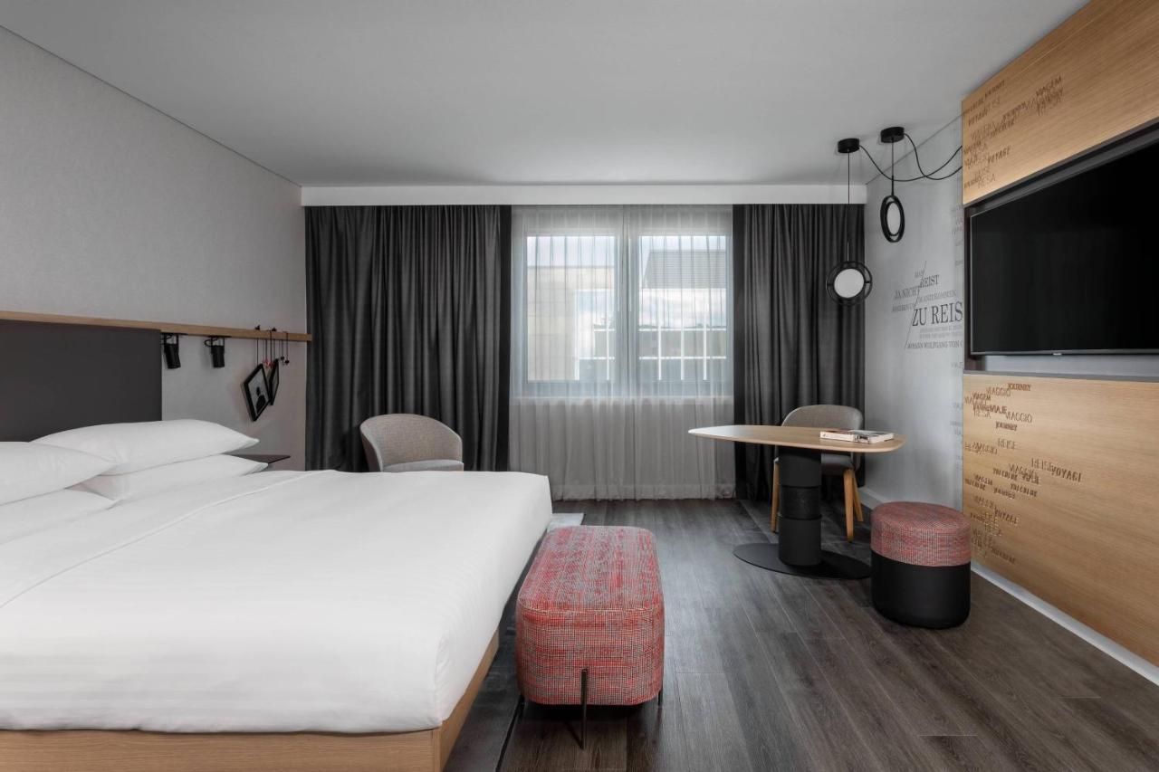 Frankfurt Airport Marriott Hotel فرانكفورت المظهر الخارجي الصورة The photo shows a modern hotel room featuring a minimalist design. There's a large bed with white linens, flanked by two bedside tables. In the corner, there are two chairs next to a small round table. A flat-screen TV is mounted on the wall opposite