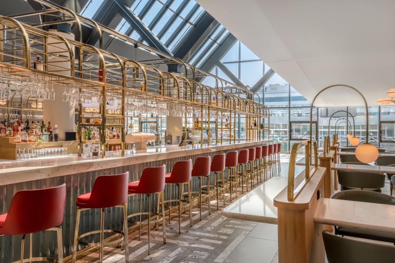 Frankfurt Airport Marriott Hotel فرانكفورت المظهر الخارجي الصورة The image shows a modern bar interior with a sleek, stylish design. There is a long bar counter made of polished materials, adorned with various drinks and glassware. High-backed red stools line the bar, offering seating for guests. The space feature