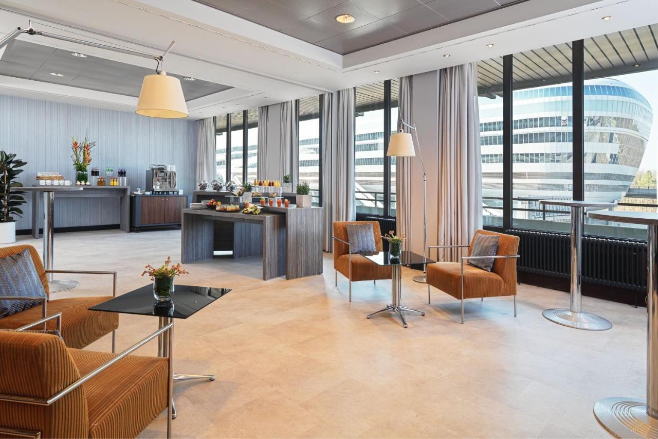 Frankfurt Airport Marriott Hotel فرانكفورت المظهر الخارجي الصورة The photo shows a modern, well-lit lounge or cafe area. There are large windows that provide a view of the outside, and the space is decorated with light-colored walls and flooring. In the foreground, there are two stylish orange armchairs and a blac