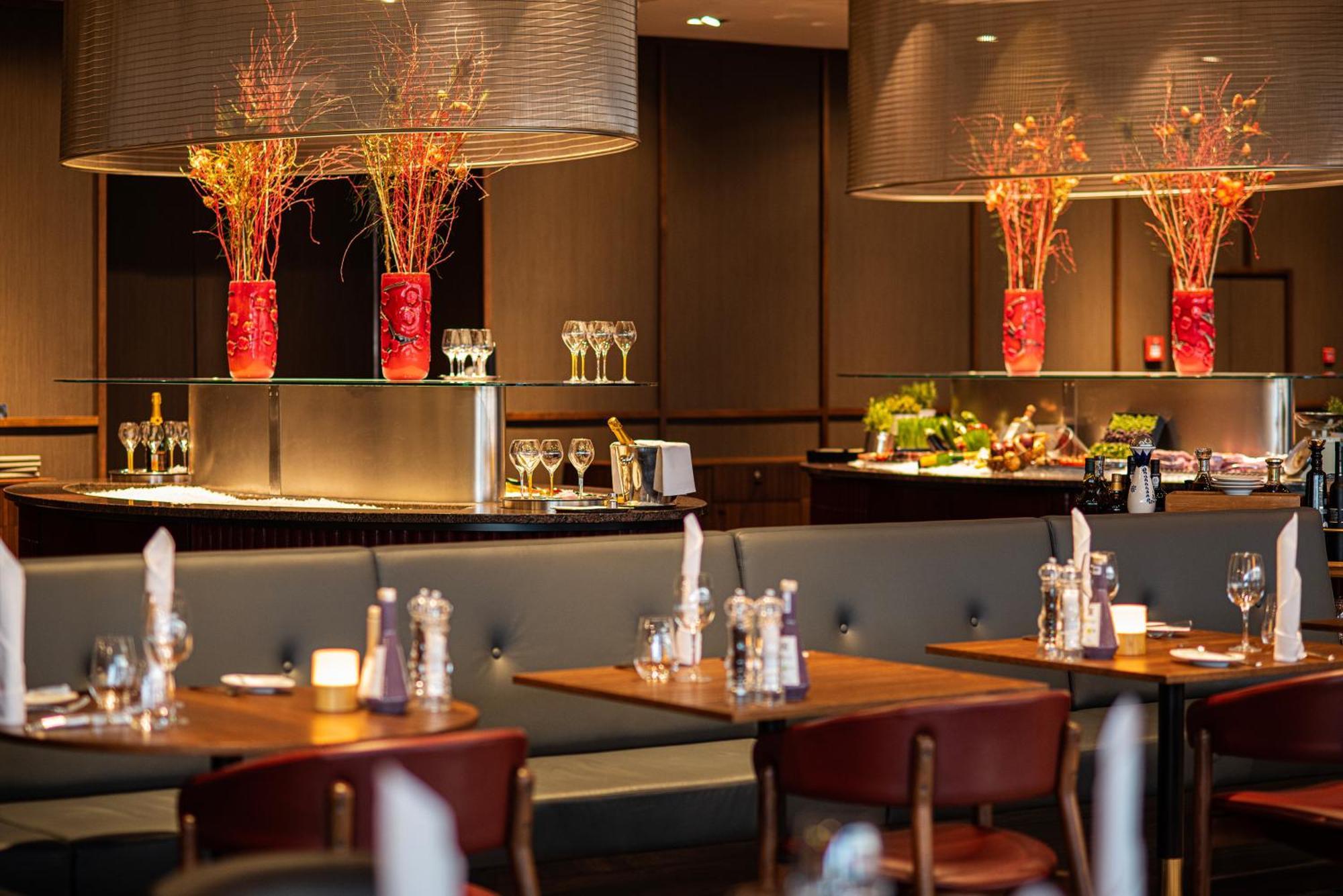 Frankfurt Airport Marriott Hotel فرانكفورت المظهر الخارجي الصورة The photo shows an elegant restaurant interior. It features wooden tables with chairs arranged neatly, creating a warm and inviting atmosphere. In the background, there are large decorative vases with colorful floral arrangements, adding a pop of col
