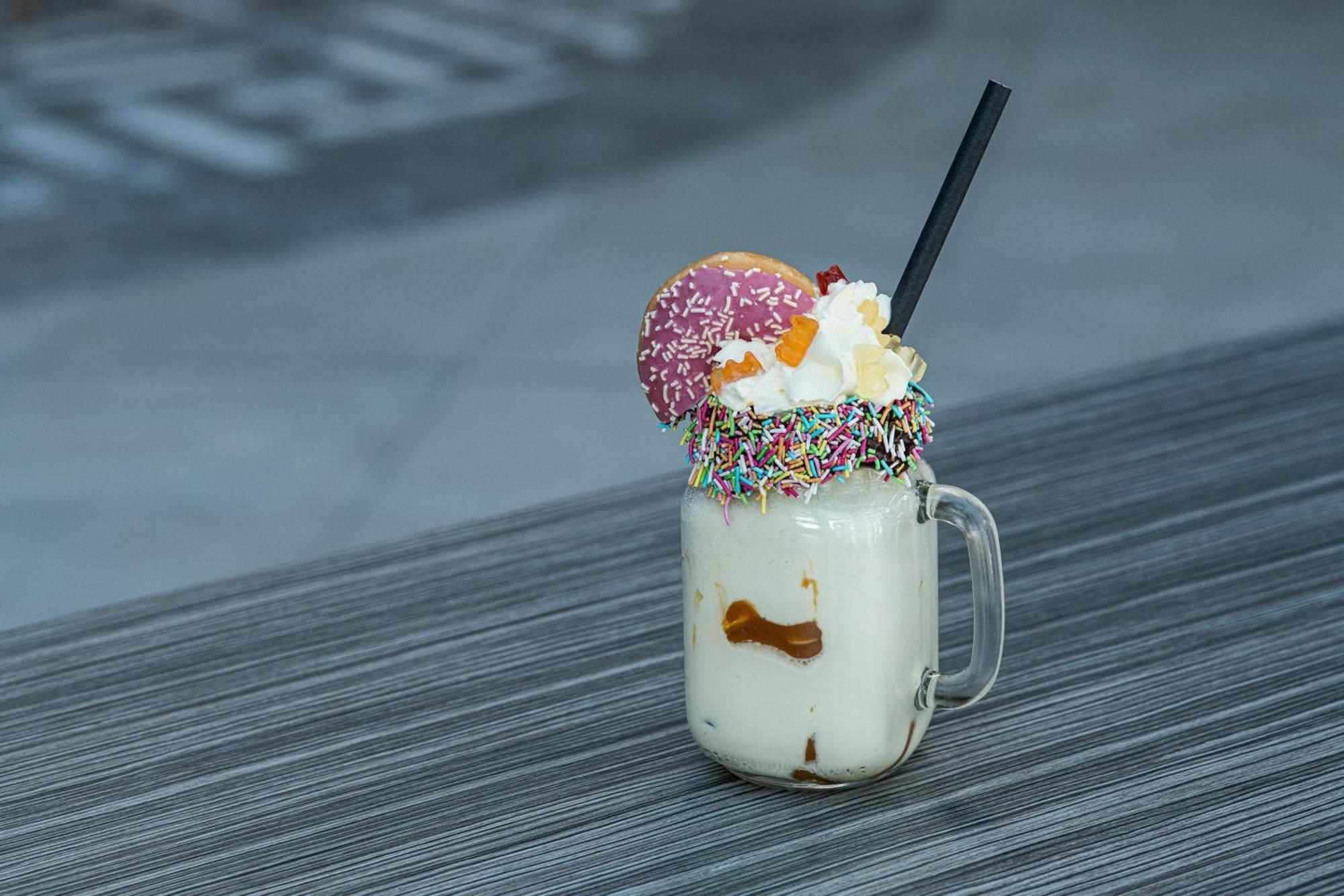 Frankfurt Airport Marriott Hotel فرانكفورت المظهر الخارجي الصورة The photo shows a large glass mug filled with a creamy beverage, likely a milkshake or dessert drink. The drink is topped with whipped cream, colorful sprinkles, and features a round donut with pink frosting and colorful sprinkles on top. A black str