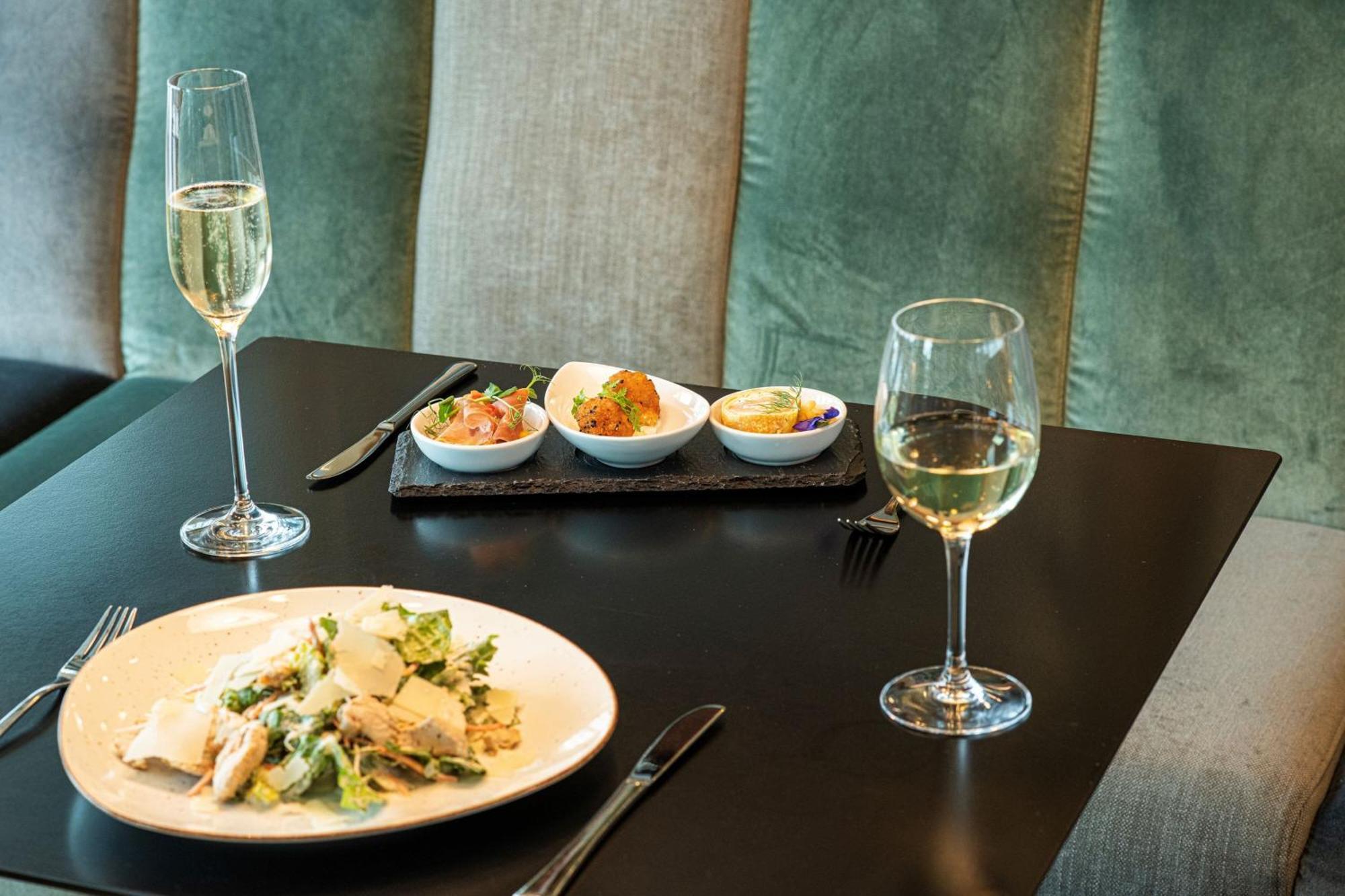 Frankfurt Airport Marriott Hotel فرانكفورت المظهر الخارجي الصورة The photo shows a dining table set for a meal. On the table, there are two glasses of sparkling wine, and a plate of salad with chicken, featuring leafy greens and possibly other ingredients. Alongside the plate, there is a small rectangular dish con