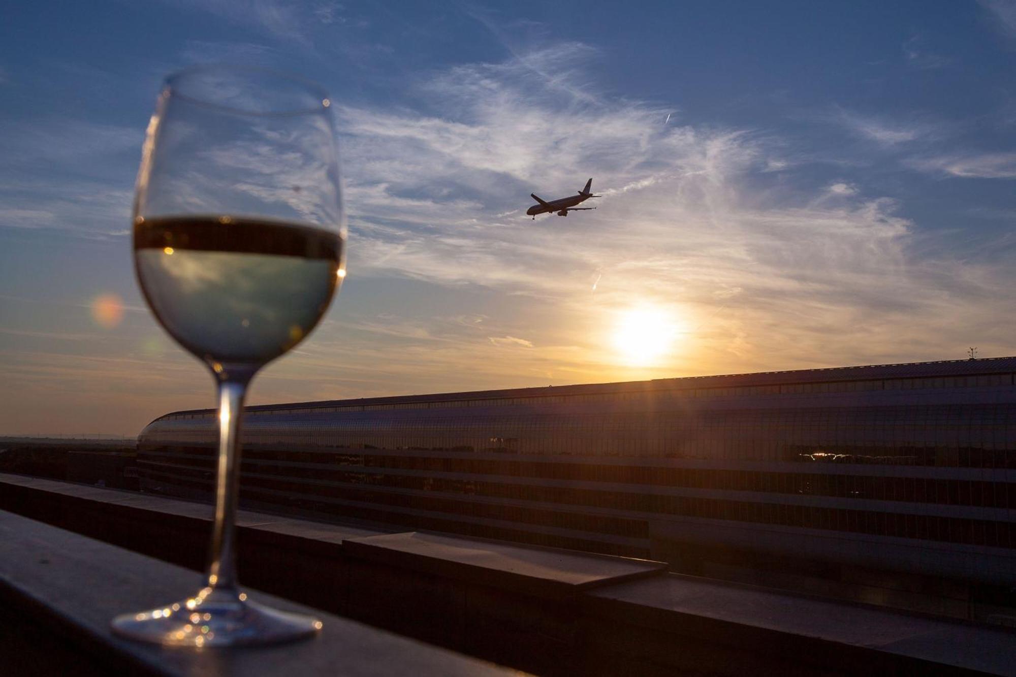 Frankfurt Airport Marriott Hotel فرانكفورت المظهر الخارجي الصورة The photo depicts a glass of white wine in the foreground, with a blurred view of an airplane flying in the sky above. The background features a beautiful sunset, casting warm colors across the clouds and sky. The setting suggests a relaxing or celeb