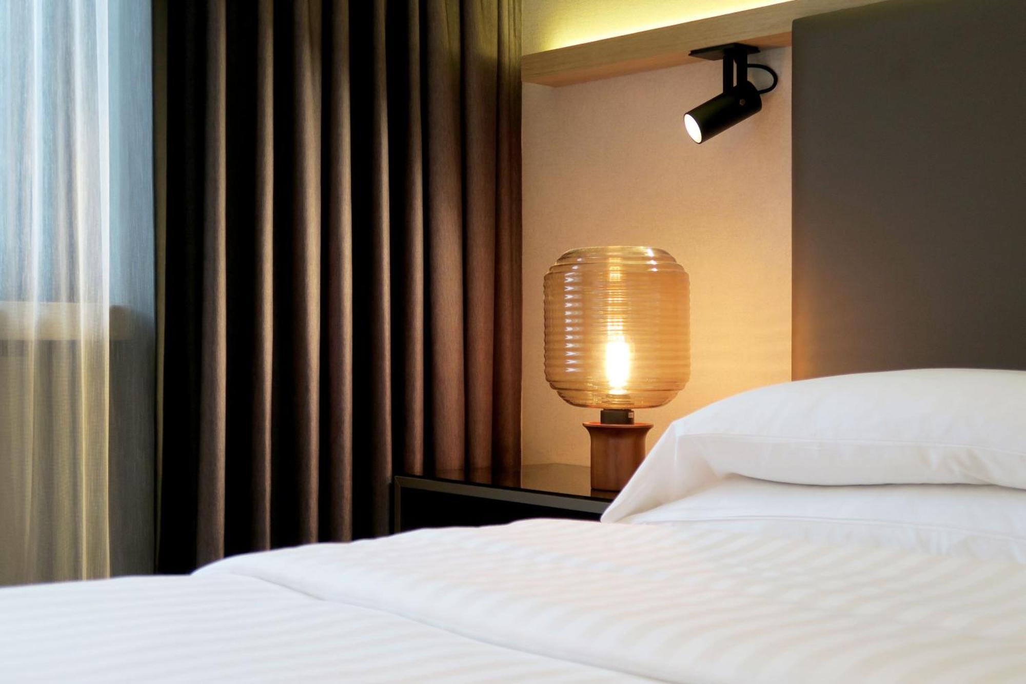 Frankfurt Airport Marriott Hotel فرانكفورت المظهر الخارجي الصورة The photo shows a serene bedroom setting. In the foreground, there is a neatly made bed with white bedding. To the side, a stylish bedside lamp with a textured glass shade emits a warm glow. Above the lamp, there is a wall-mounted light fixture that 