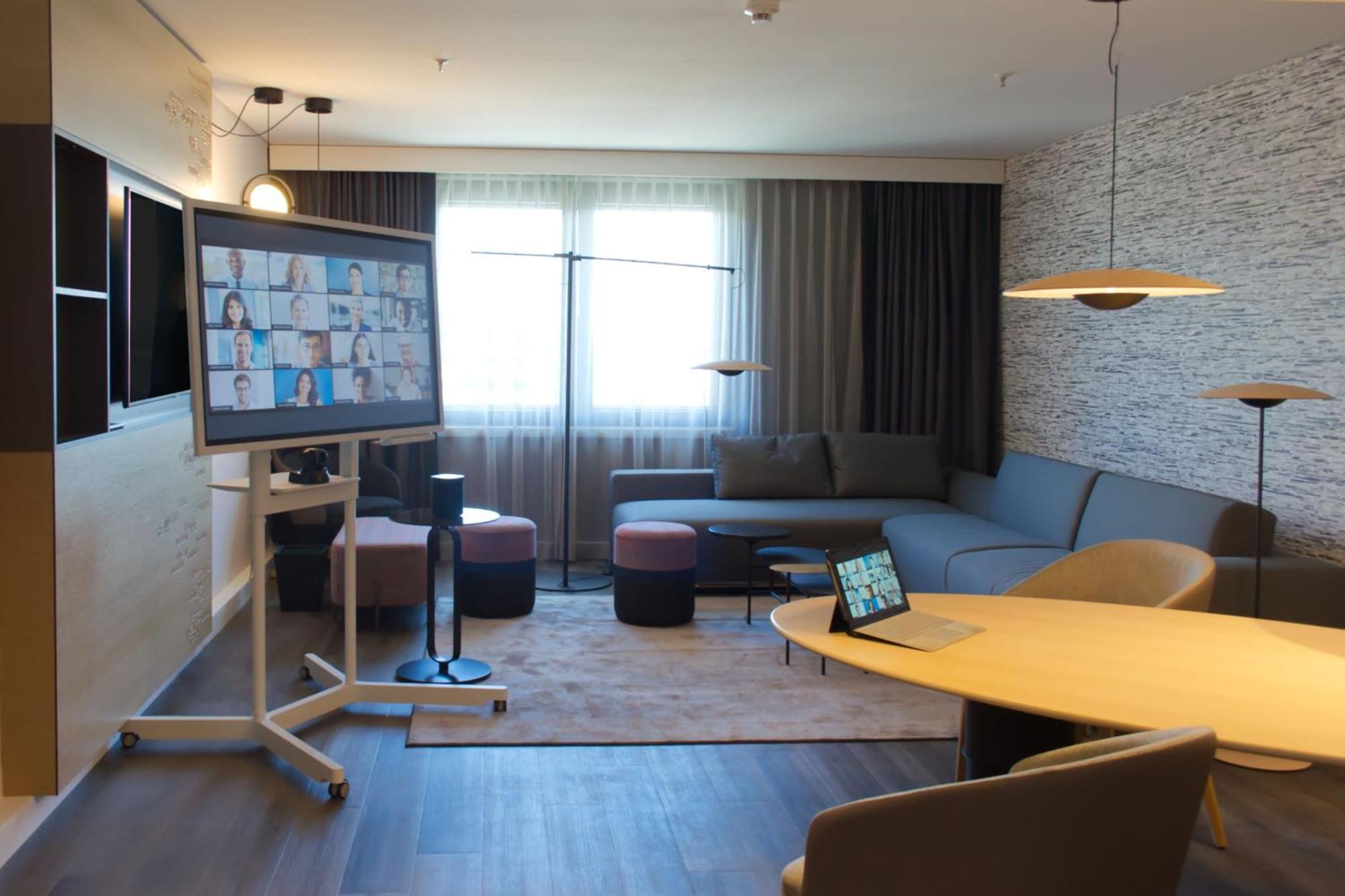Frankfurt Airport Marriott Hotel فرانكفورت المظهر الخارجي الصورة The image shows a modern meeting room or office space designed for collaboration. It features a large screen displaying video conference participants, indicating that it's equipped for virtual meetings. There is a comfortable seating area with a gray