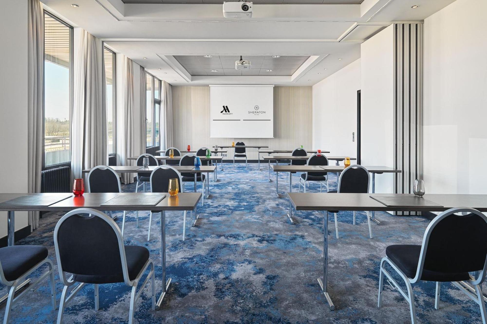 Frankfurt Airport Marriott Hotel فرانكفورت المظهر الخارجي الصورة The photo shows a spacious meeting room designed for events or conferences. There are several tables arranged in a classroom style, with chairs positioned around them. The decor includes a modern carpet with a blue and white design. At the front, the