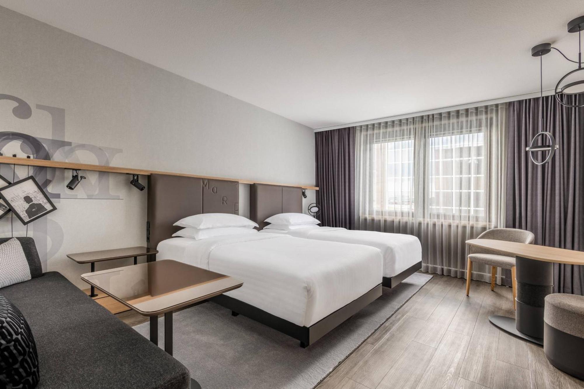 Frankfurt Airport Marriott Hotel فرانكفورت المظهر الخارجي الصورة The photo shows a modern hotel room featuring two beds with white linens and dark-colored headboards. There is a small table beside each bed, and a sofa is visible against the wall. Large windows with sheer curtains allow natural light to brighten th