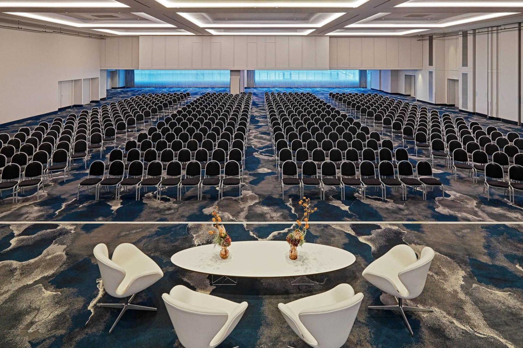 Frankfurt Airport Marriott Hotel فرانكفورت المظهر الخارجي الصورة The photo shows a large conference or event room set up for seating. It features rows of black chairs arranged in a theater style across a blue and white patterned carpet. In the foreground, there is a stylish white circular table accompanied by two 