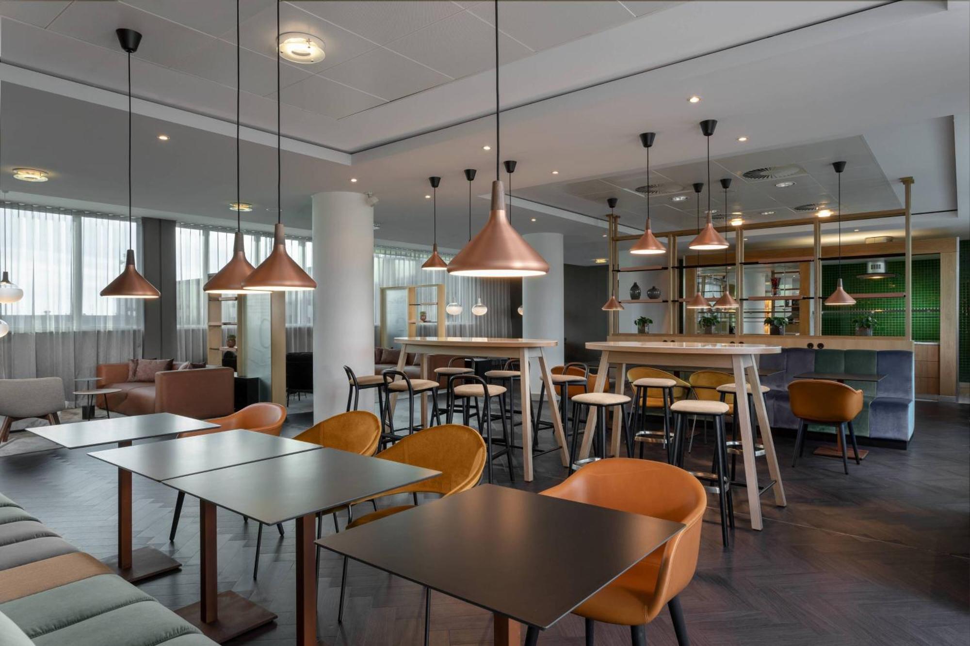Frankfurt Airport Marriott Hotel فرانكفورت المظهر الخارجي الصورة The photo shows the interior of a modern café or lounge. It features a mix of seating arrangements, including tall tables with barstools and lower tables with orange chairs. The space is well-lit, with stylish pendant lights hanging from the ceiling.