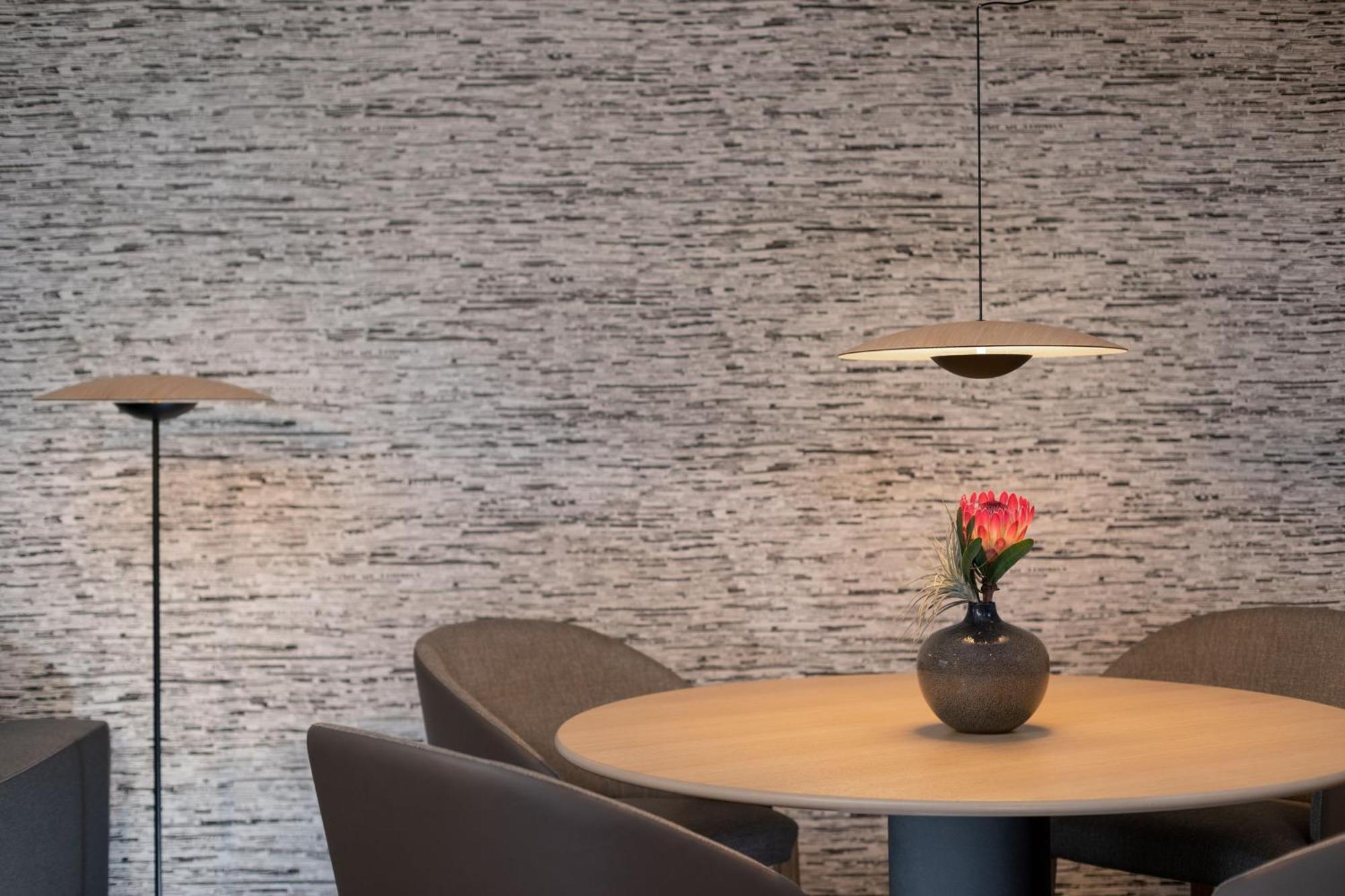 Frankfurt Airport Marriott Hotel فرانكفورت المظهر الخارجي الصورة The photo features a modern interior scene. In the center, there is a round wooden table surrounded by four padded chairs. At the table's center, a vase holds a single flower with a pink bloom. Above the table, a contemporary pendant light provides s
