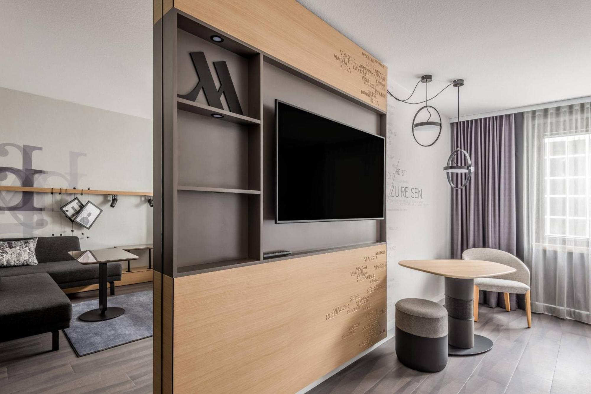 Frankfurt Airport Marriott Hotel فرانكفورت المظهر الخارجي الصورة The photo shows a contemporary hotel room or living space. It features a wooden accent wall with built-in shelves, containing decorative items and a television set. There is a small dining table with two upholstered stools nearby. The space is well-l