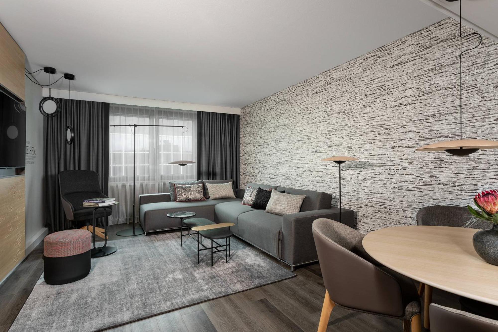 Frankfurt Airport Marriott Hotel فرانكفورت المظهر الخارجي الصورة The photo shows a modern living room or lounge area that features a stylish and minimalist design. There is a comfortable gray sofa with various cushions arranged on it. Next to the sofa, there's a small coffee table and a few additional accent table