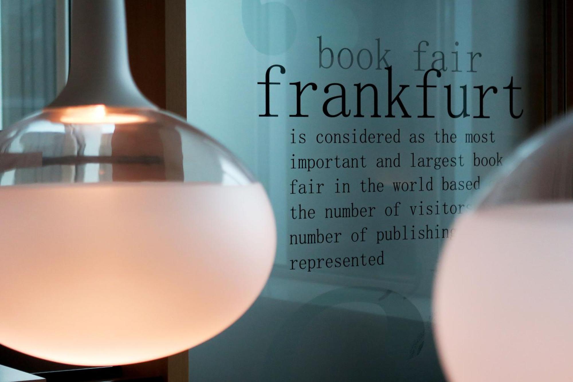 Frankfurt Airport Marriott Hotel فرانكفورت المظهر الخارجي الصورة The photo features two modern pendant light fixtures hanging from the ceiling. In the background, there is a piece of glass or wall with text that reads about the Frankfurt Book Fair, stating that it is the most important and largest book fair in the