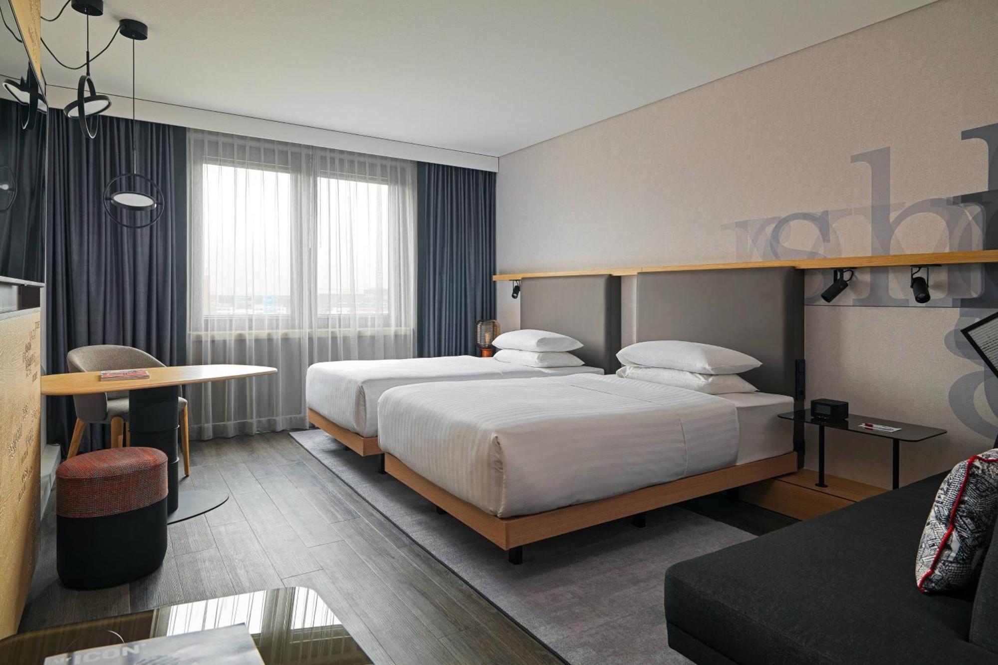 Frankfurt Airport Marriott Hotel فرانكفورت المظهر الخارجي الصورة The photo shows a modern hotel room featuring two twin beds with white linens positioned against a wooden headboard. There are stylish bedside lamps attached to the headboards. The room has large windows with sheer white curtains that let in natural 