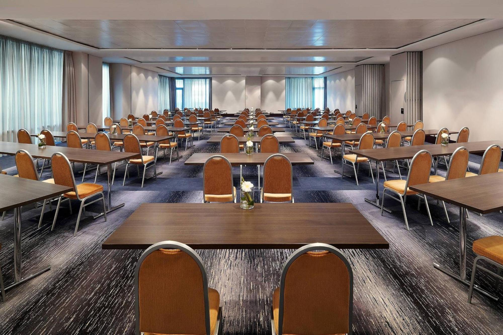 Frankfurt Airport Marriott Hotel فرانكفورت المظهر الخارجي الصورة The photo shows a large conference room arranged with multiple rows of tables and chairs. The setup is designed for a gathering, such as a seminar or workshop. Each table has a small decorative item, possibly a flower or a plant, at its center. The r