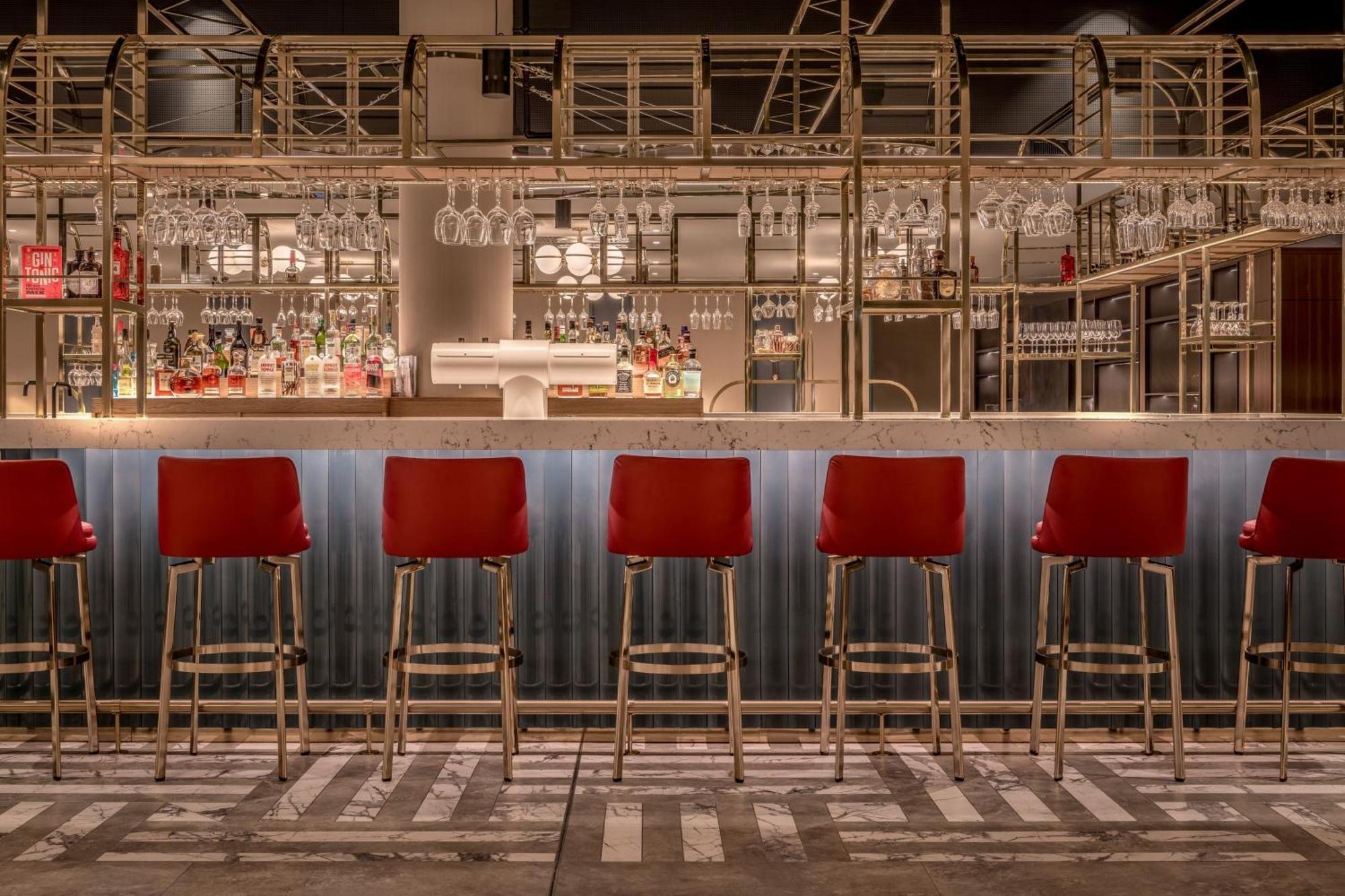 Frankfurt Airport Marriott Hotel فرانكفورت المظهر الخارجي الصورة The photo shows a stylish bar area with a row of red bar stools positioned in front of a long counter. The bar has a modern design, featuring a combination of metal and wood elements. Behind the bar, there are shelves holding various bottles of alcoh