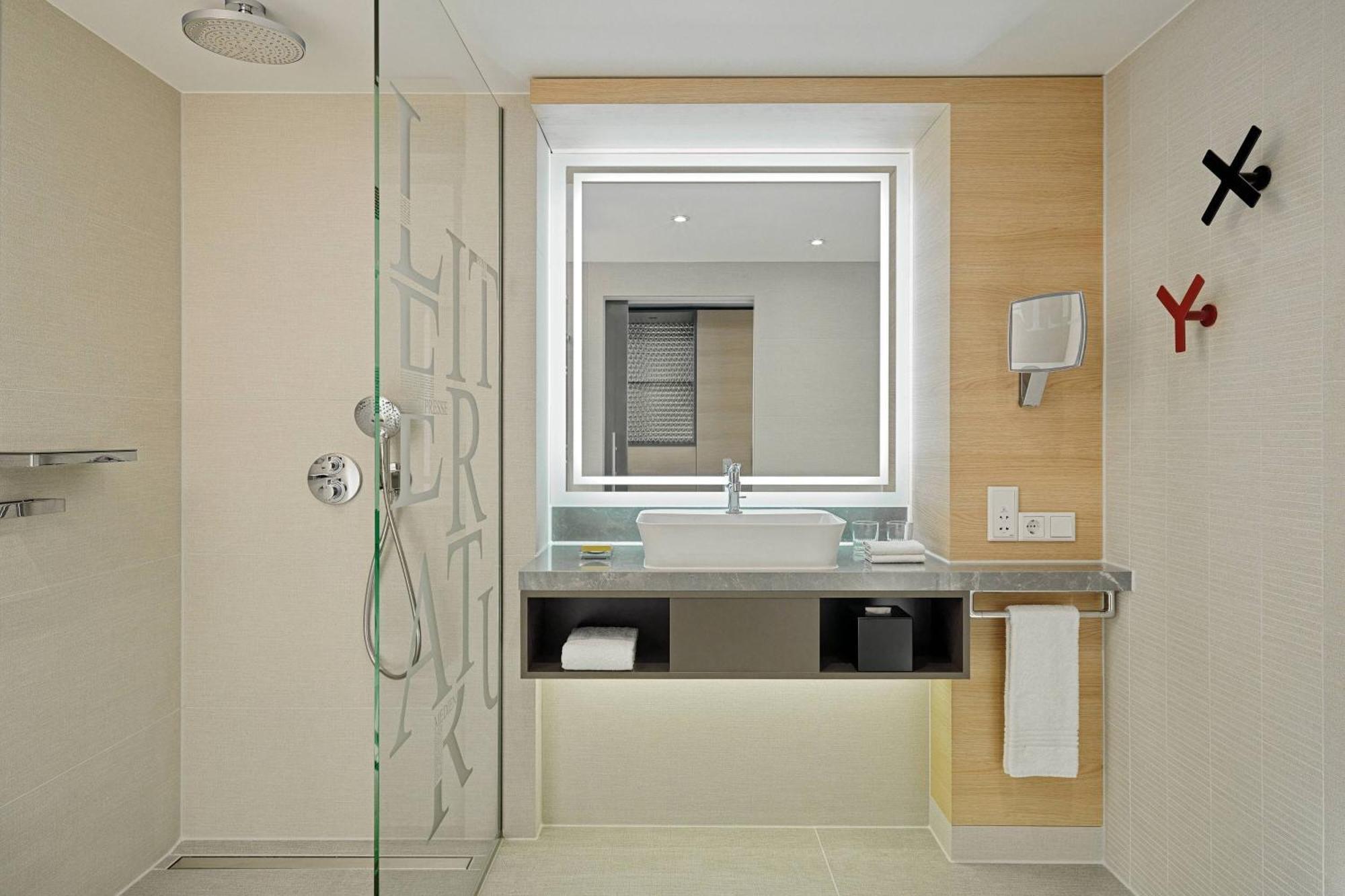 Frankfurt Airport Marriott Hotel فرانكفورت المظهر الخارجي الصورة The photo shows a modern bathroom with a sleek design. It features a glass shower enclosure on the left, a stylish sink with a large mirror above it, and a countertop underneath the mirror. The wall behind the sink is made of light-colored wood, enha