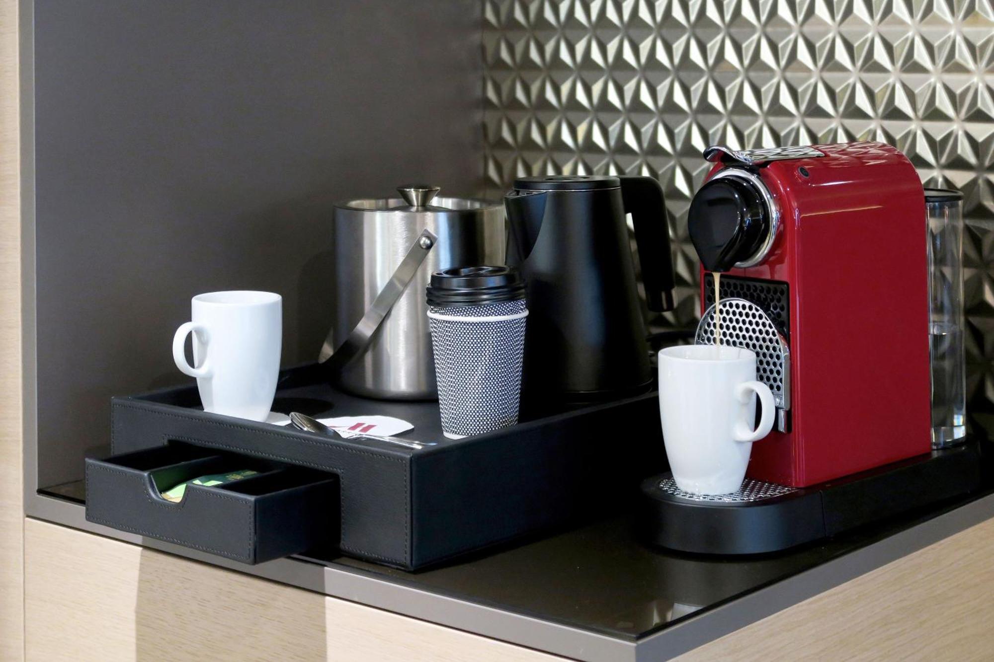 Frankfurt Airport Marriott Hotel فرانكفورت المظهر الخارجي الصورة The photo shows a coffee station setup. It includes a red coffee machine on the right, designed for making espresso. To the left of the machine, there are two white cups and a silver coffee pot, along with a black container that seems to hold some it