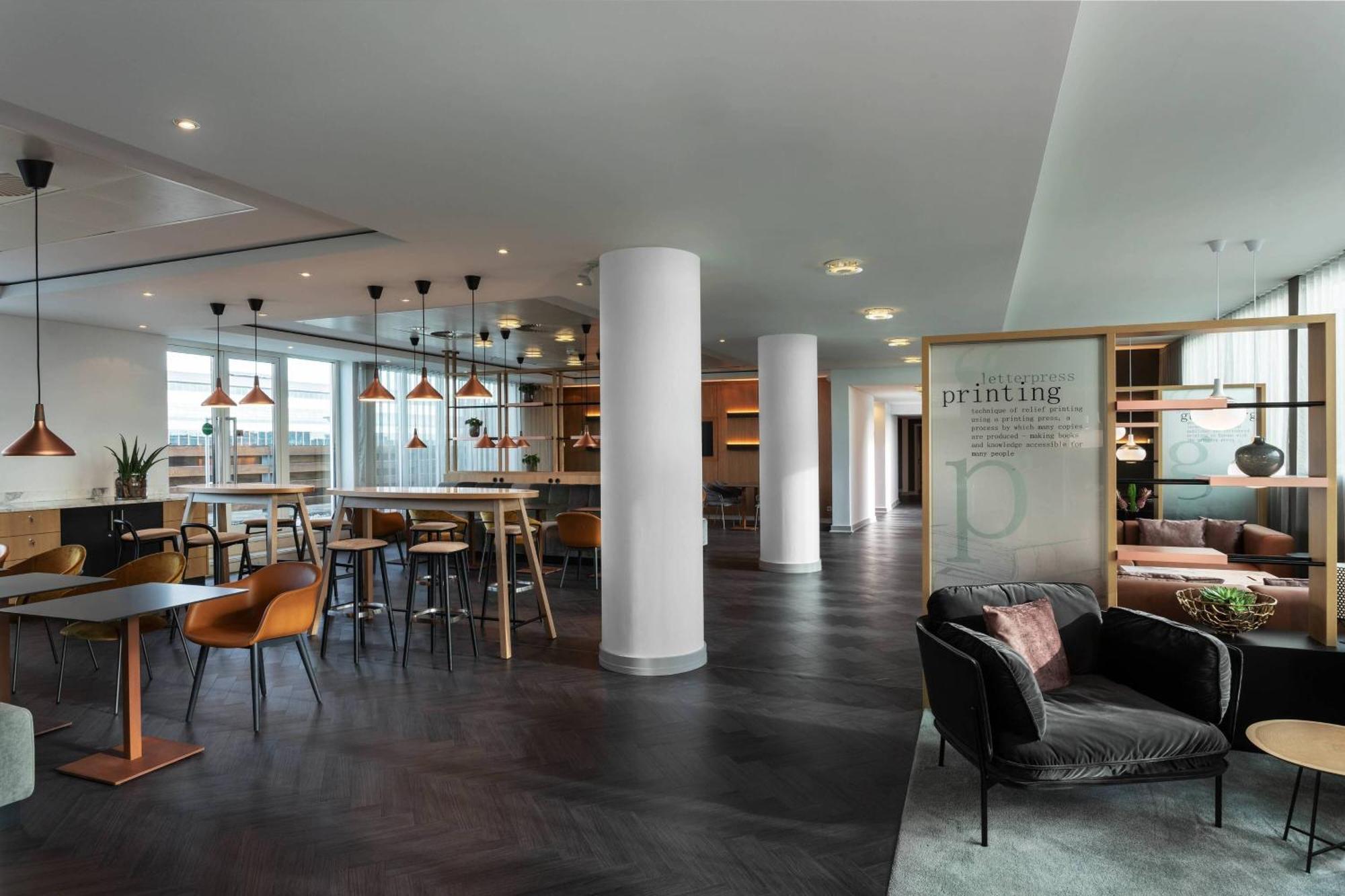 Frankfurt Airport Marriott Hotel فرانكفورت المظهر الخارجي الصورة The photo shows a modern, stylish interior of a workspace or a lounge area. It features a mix of seating arrangements, including a cozy dark sofa and tables with chairs. The space has an open layout with sleek flooring, large windows allowing natural