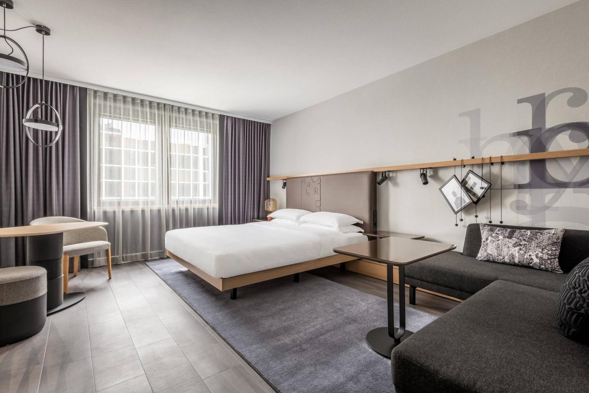 Frankfurt Airport Marriott Hotel فرانكفورت المظهر الخارجي الصورة The photo shows a modern hotel room featuring a bed with white linens, a stylish wooden headboard, and a nightstand. There's a comfortable seating area with a dark sofa and a side table. A small dining table with two chairs is also visible. The windo