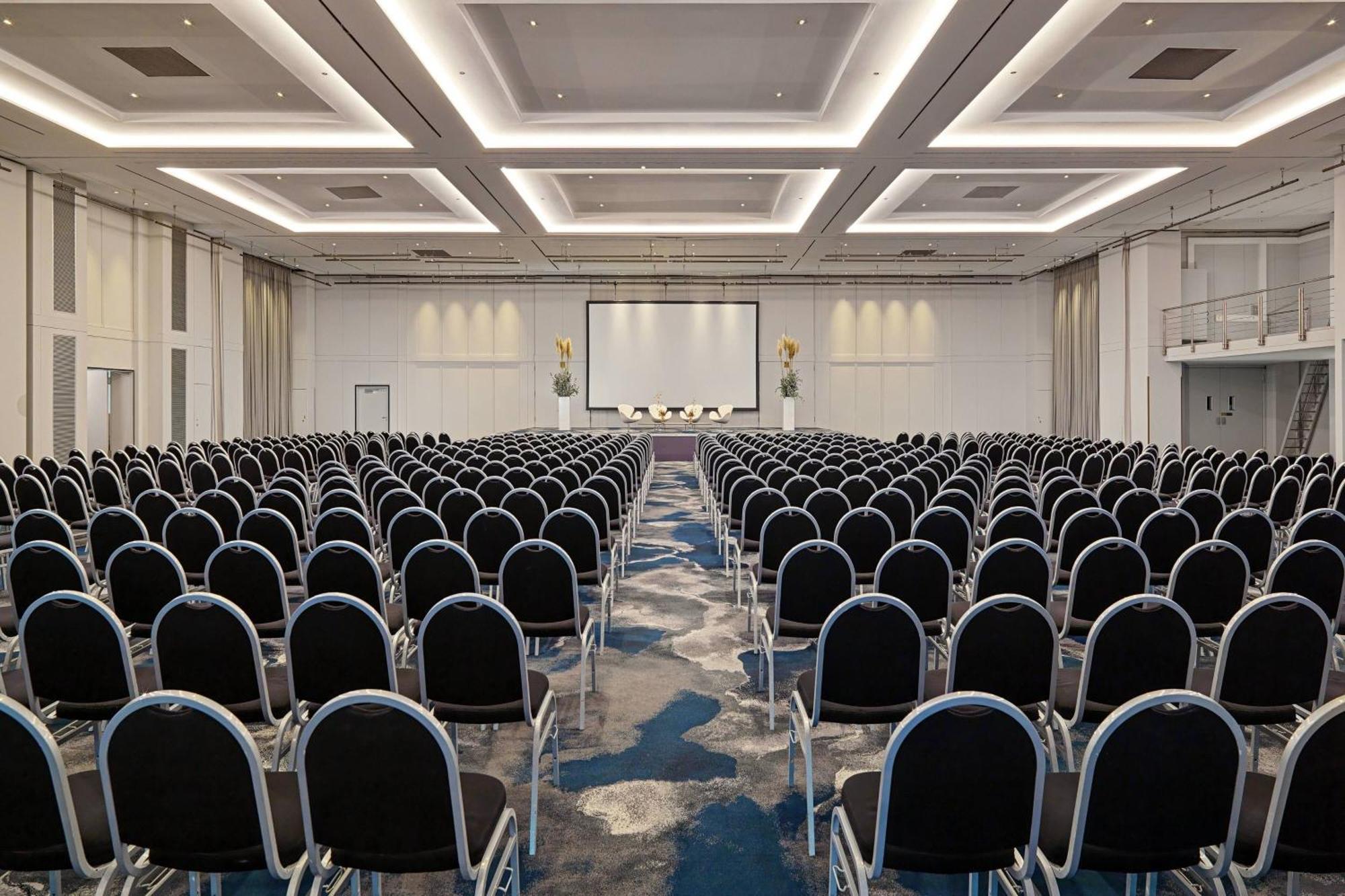 Frankfurt Airport Marriott Hotel فرانكفورت المظهر الخارجي الصورة The photo shows a large conference room arranged for a presentation or event. The space is filled with rows of black chairs, all facing a central stage area where a large screen is positioned. The room features modern lighting and a clean, profession